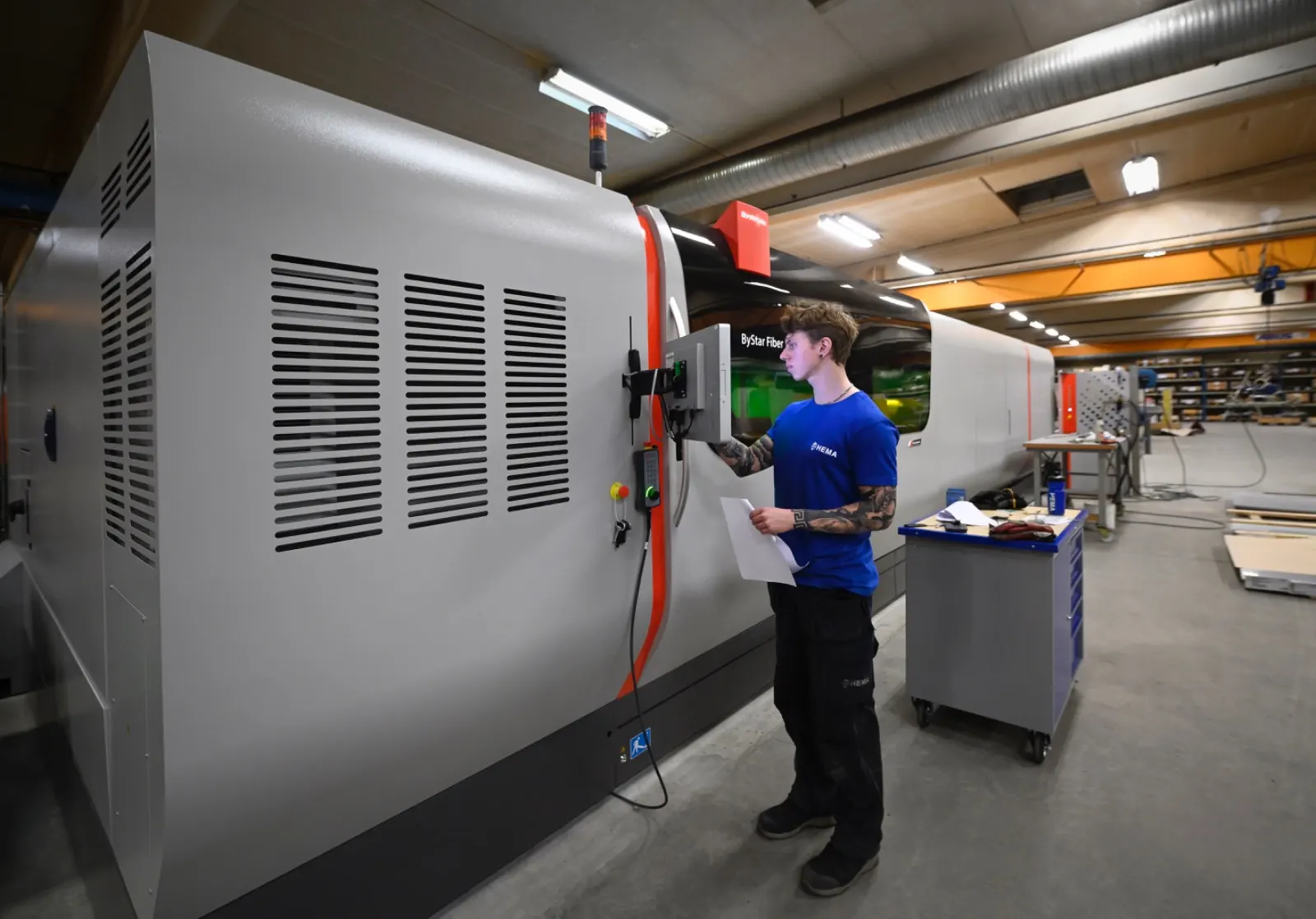 Operatör i HEMA-tröja vid laserskärmaskinens manöverpanel, med underlaget i handen.